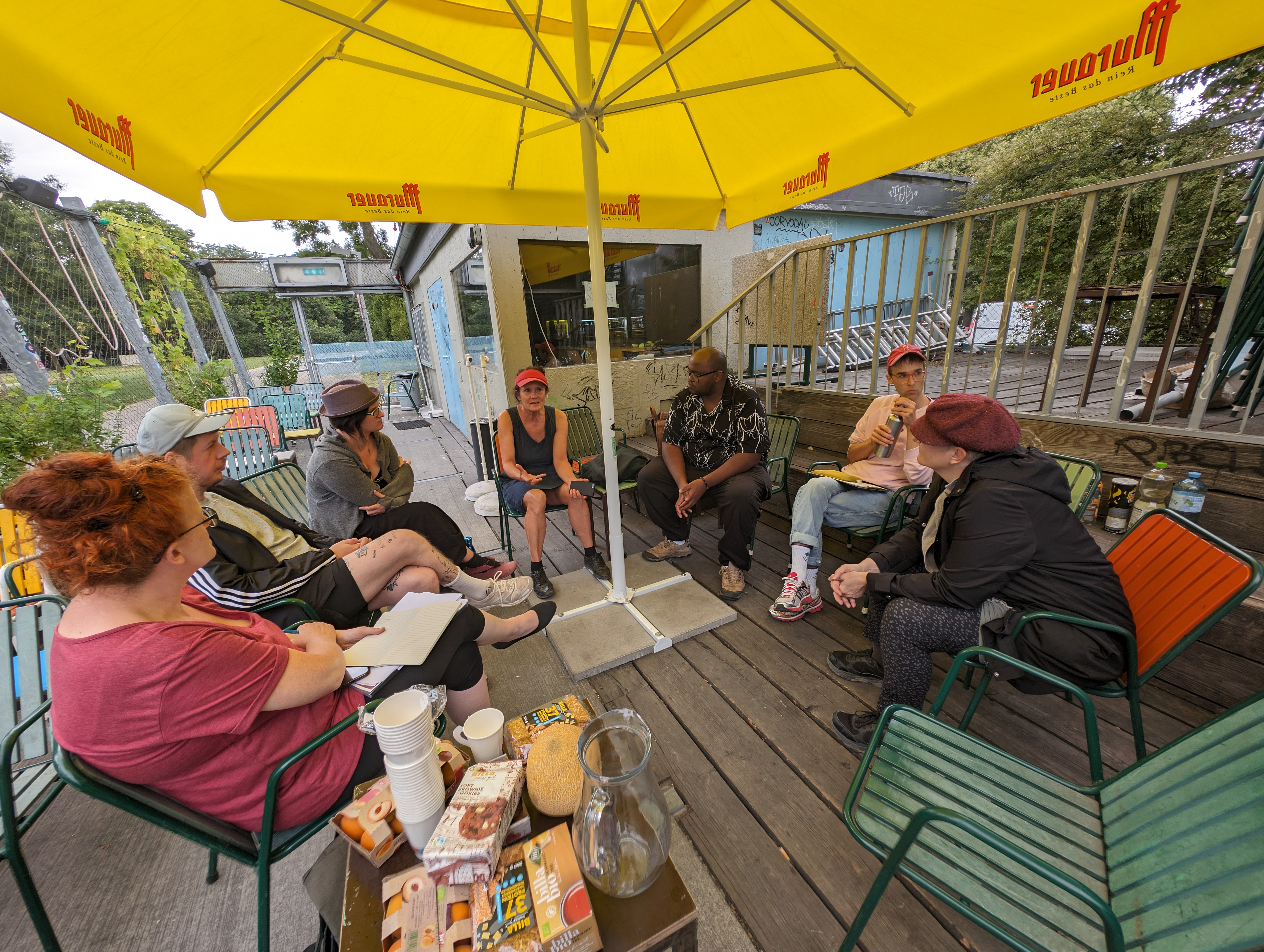 Foto: Susanne Rogenhofer Teilnehmende, die im Kreis unter einem Sonnenschirm sitzen und diskutieren