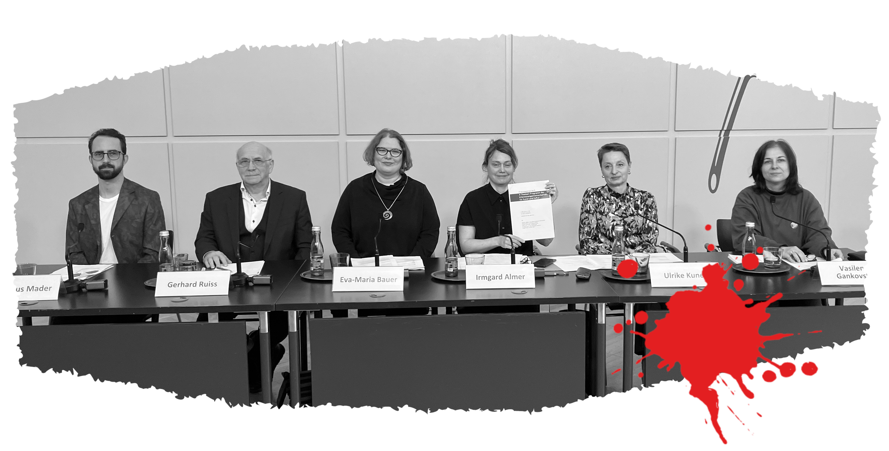 6 Personen sitzen an einem Podium, eine von ihnen hält die Presseunterlagen hoch.