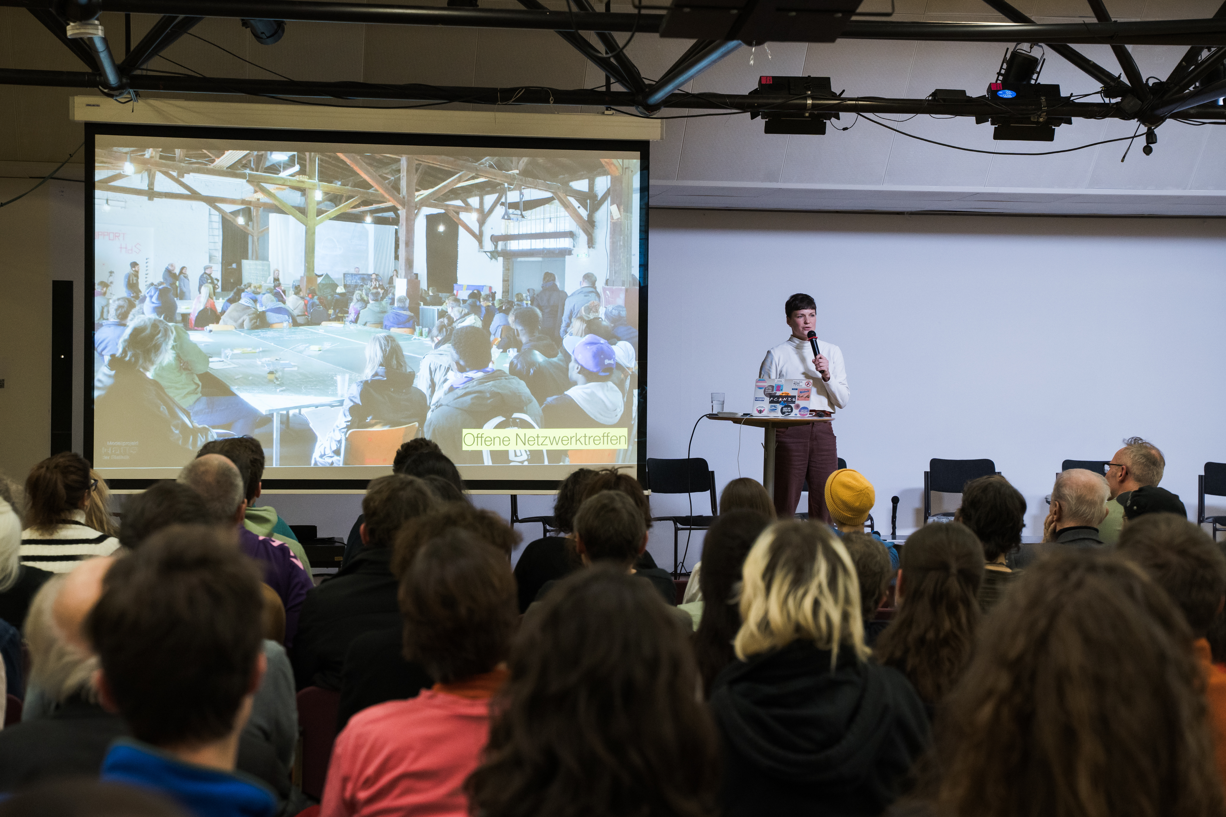 Foto: eSeL.at/Sarah Tasha Publikum vor einer Leinwand, auf der Personen sitzen, großteil an einem Tisch, neben der Leinwand spricht eine Person in ein Mikrofon